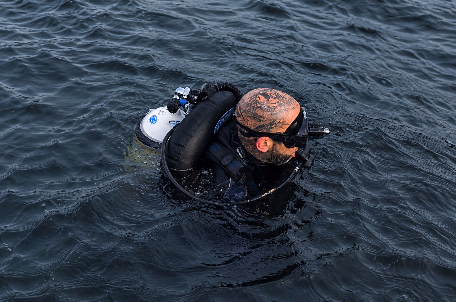 Tattooed scuba diver wearing a black wetsuit and mask floating on dark water surface.